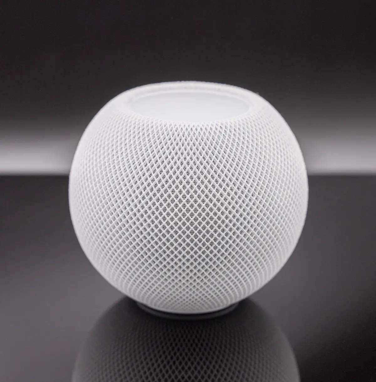 Apple HomePod mini smart speaker in white, viewed from the front on a reflective surface